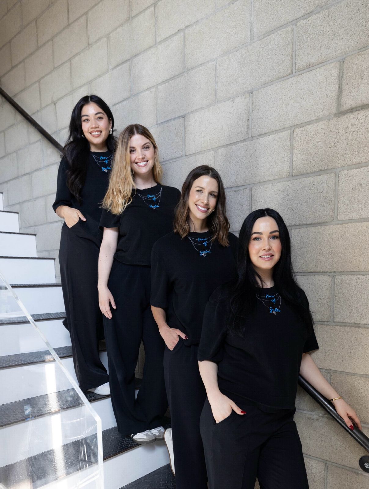 Four women in black outfits smile on indoor stairs by a concrete wall, showing off their best Face of Jules facial glow.