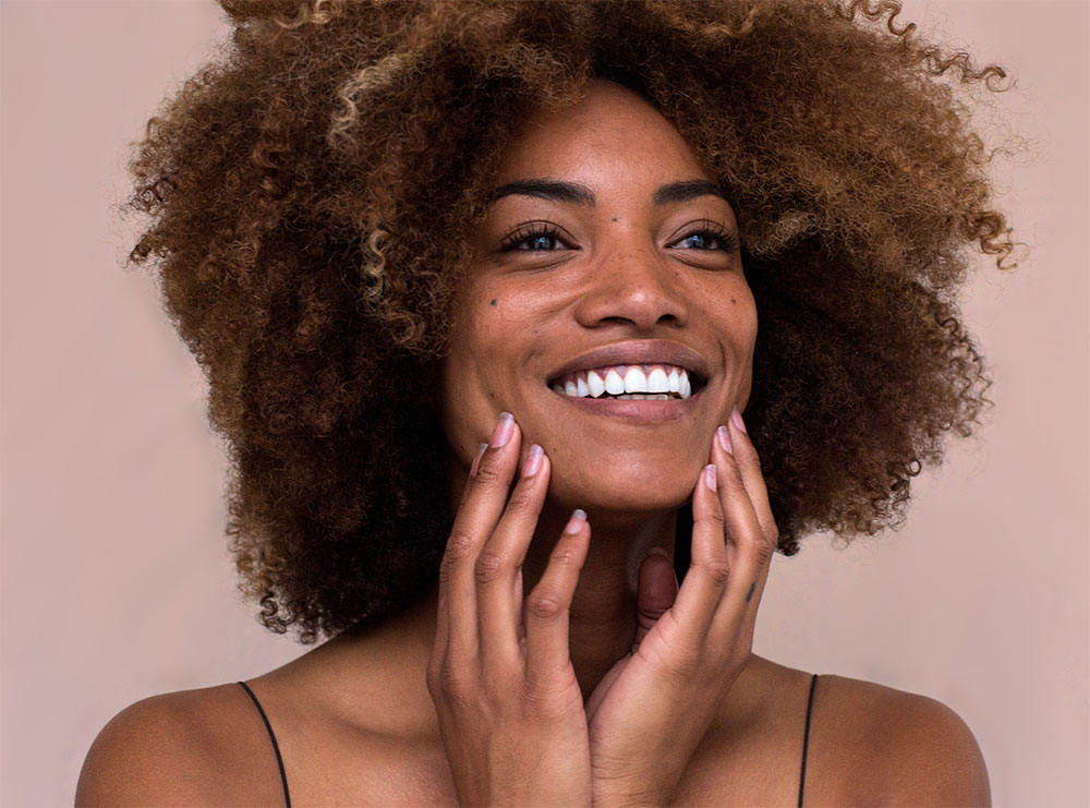 Smiling woman with curly hair touches her face with both hands, showcasing her best facial features against a light background.