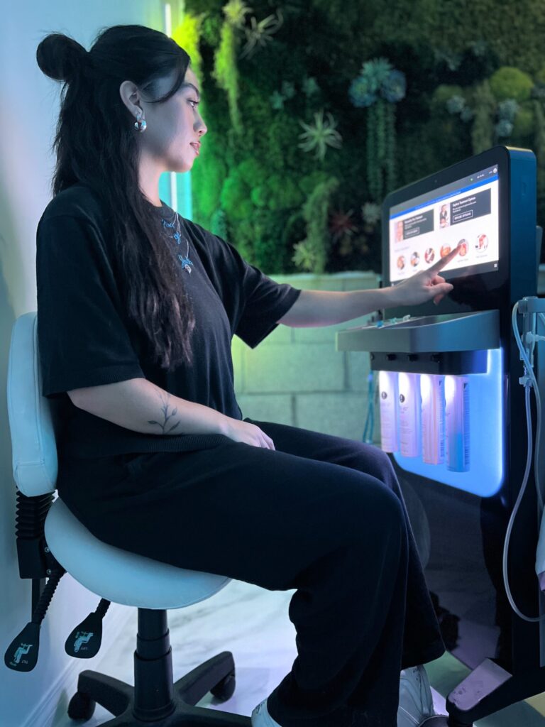 A woman sits on a stool using a touchscreen device in a modern, plant-decorated Los Angeles room.
