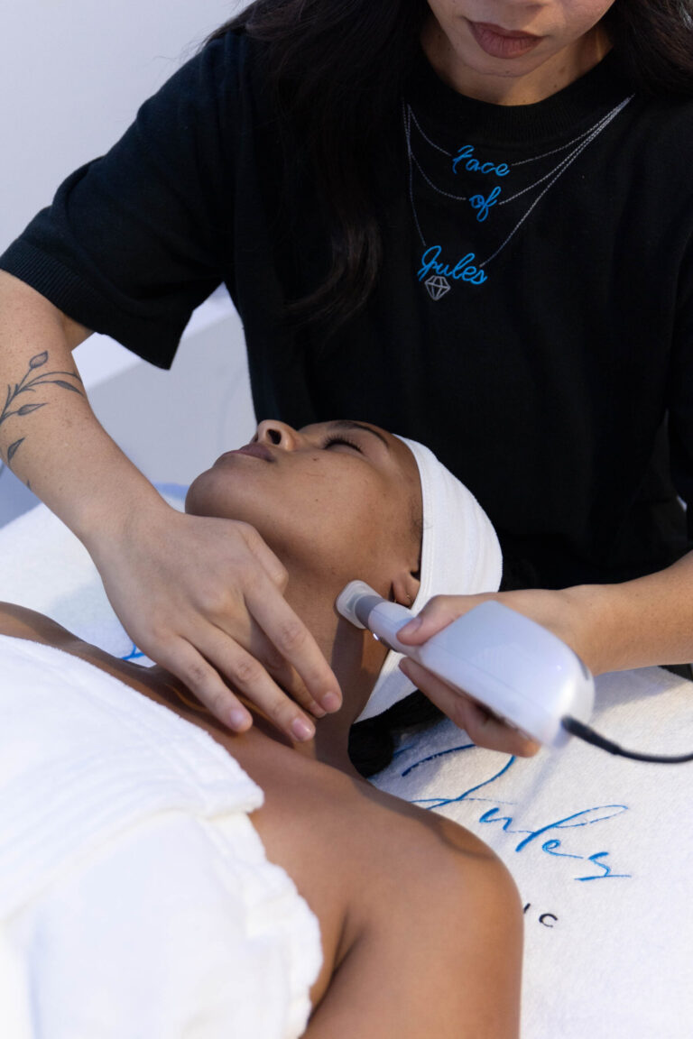 A woman enjoys the best facial at Face of Jules, relaxing on a towel-covered bed with a handheld device treatment.