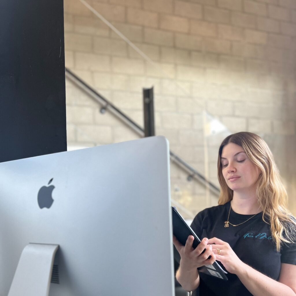 Woman standing at a desk using a tablet in front of an Apple computer, with a modern Hollywood office background.
